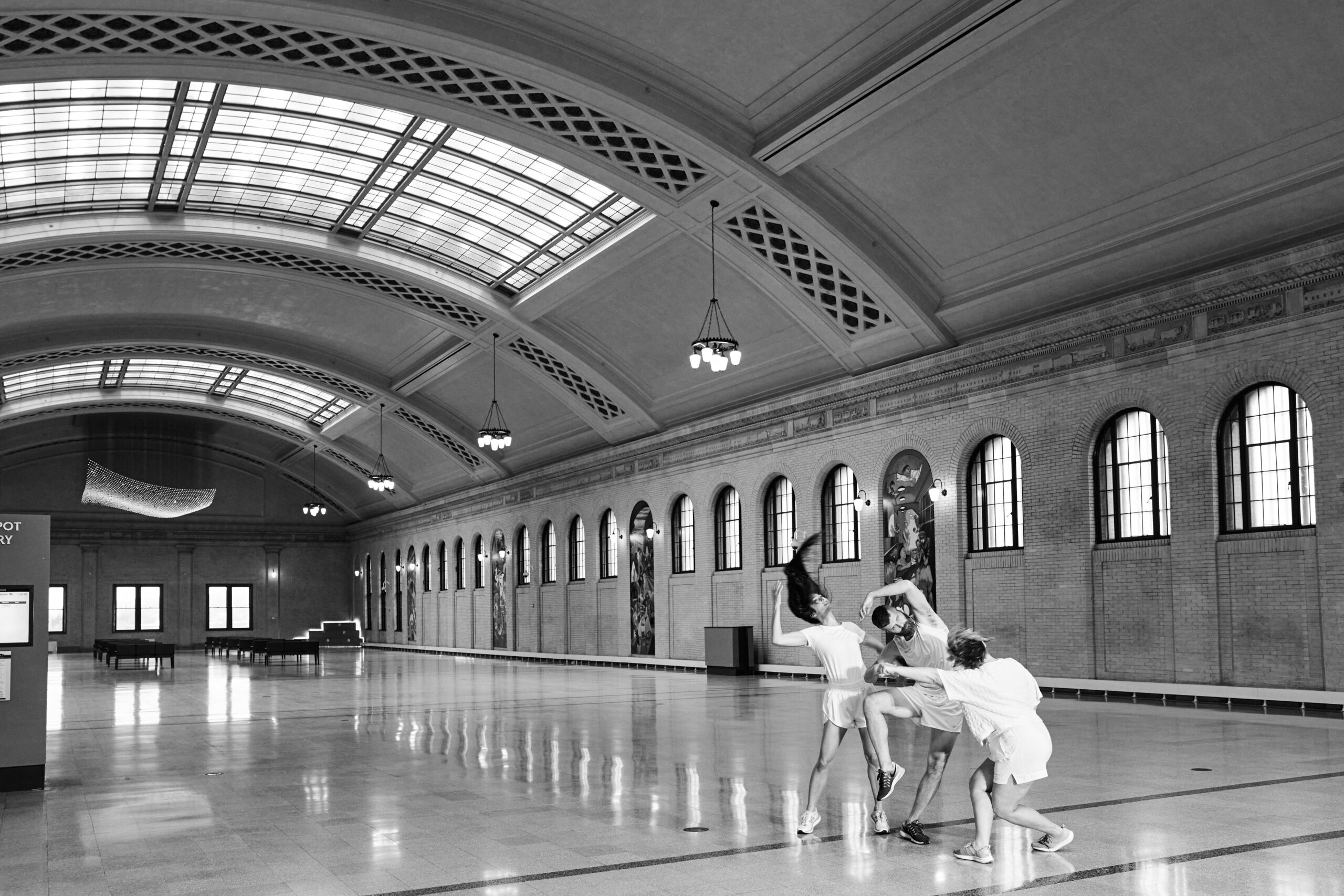 Three dancers toss their limbs in a tight group in the midst of a large, arched indoor space
