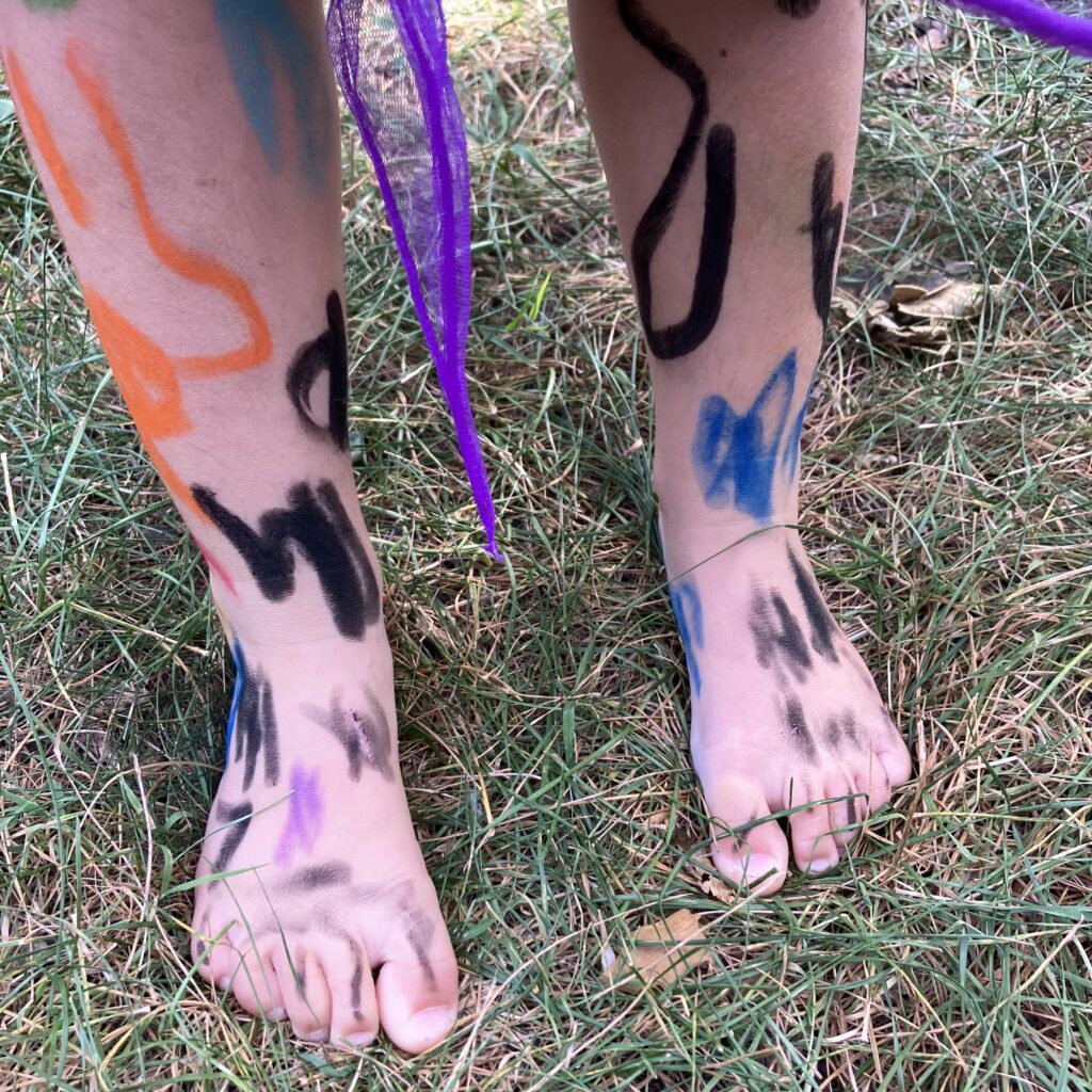 A young person's feet and legs covered in black, orange, blue and purple scrawls, stands in grass