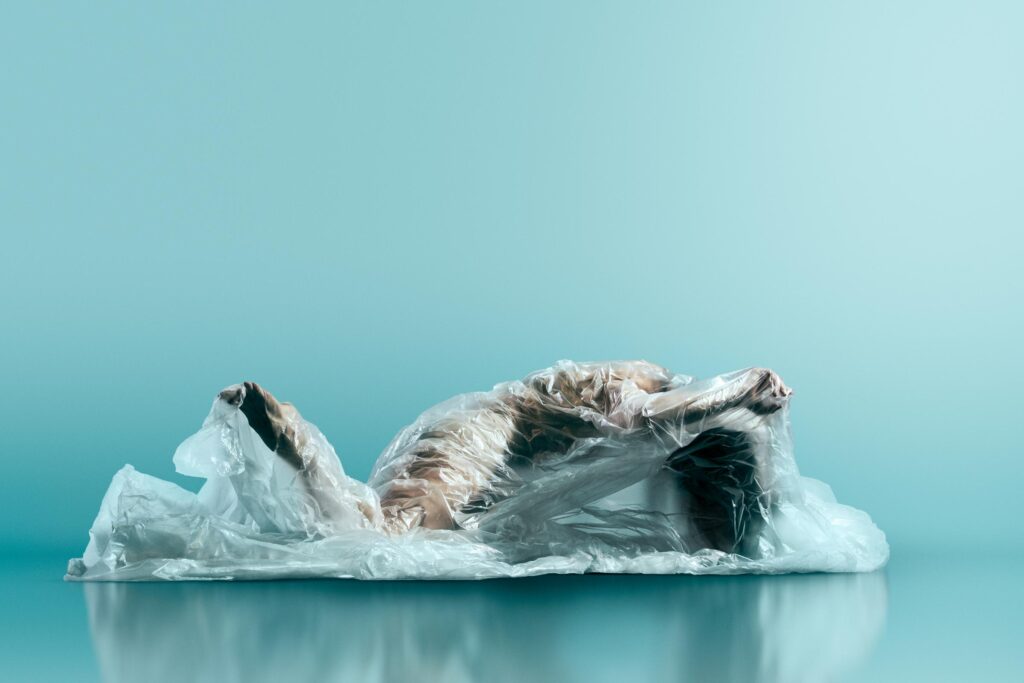 A dancer with light skin lies underneath a sheet of clear plastic.