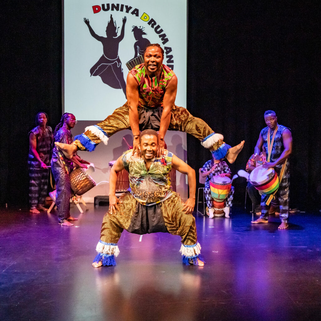 One dancer jumps over the other with drummer playing in the backround