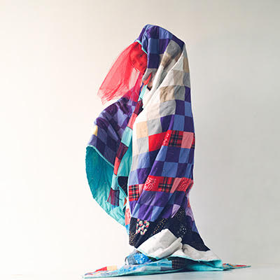 Emily Johnson, obscured by a draped colorful quilt, stands in front of a neutral backdrop.